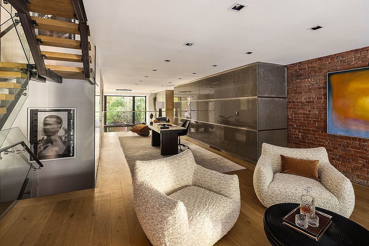 This interior shot showcases a modern living room with hardwood floors, exposed brick, and contemporary furnishings. The room features two textured armchairs, a black coffee table, and a large abstract painting, creating a stylish and inviting atmosphere. A staircase with glass railings adds a touch of elegance, while a long desk and storage unit suggest a versatile living and workspace.