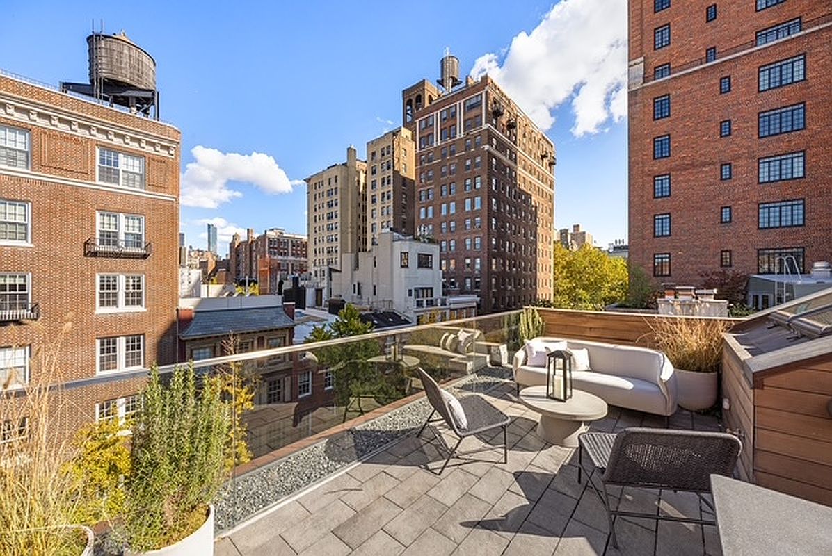This image showcases a stylish rooftop patio or balcony in an urban setting. The space is furnished with modern outdoor furniture, including a sofa, chairs, and a coffee table, creating an inviting lounge area. A glass railing provides unobstructed views of the surrounding cityscape, featuring brick buildings and water towers, enhancing the property's appeal.