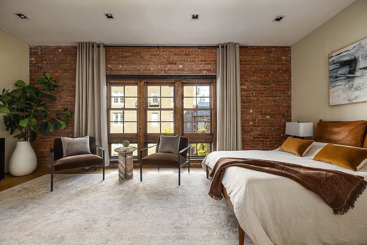 This is a primary bedroom featuring exposed brick walls, large windows, and neutral-toned decor. The room includes a bed with a white duvet and brown throw, two armchairs, and a side table. The overall style is modern and industrial, creating a cozy and inviting atmosphere.