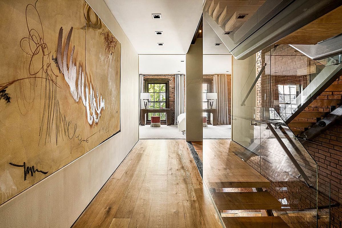 This interior shot showcases a modern hallway with hardwood flooring leading to a bedroom area, featuring a large abstract art piece on the left wall. To the right, a staircase with glass railings and exposed brick adds an industrial-chic touch. The hallway is well-lit with recessed lighting, creating a bright and inviting atmosphere.