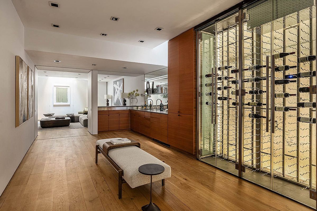 This interior shot showcases a modern hallway with light wood flooring and a built-in wine cellar with glass doors. The hallway leads to a living area, visible through a doorway, and features a sleek bench with a small side table. The overall design is minimalist and sophisticated, emphasizing clean lines and natural materials.