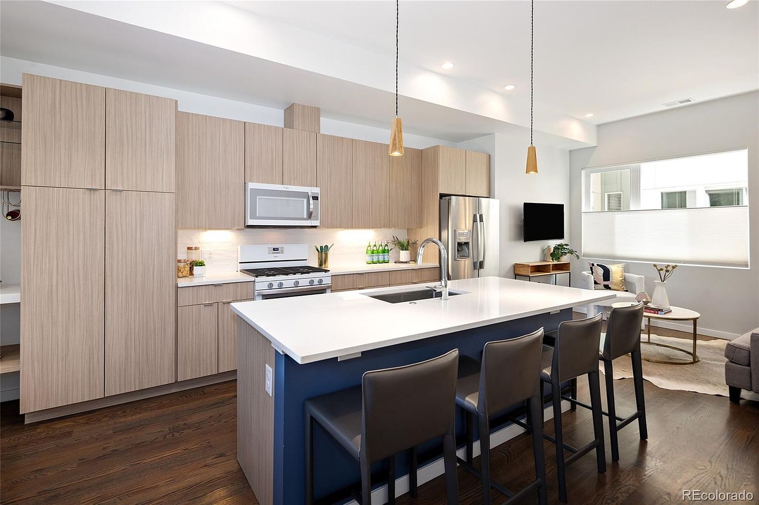 This is a well-lit, modern kitchen featuring sleek, light wood cabinetry and a large island with a white countertop and blue base. Stainless steel appliances, including a microwave and refrigerator, complement the contemporary design. The kitchen seamlessly transitions into a living area, creating an open and inviting space.