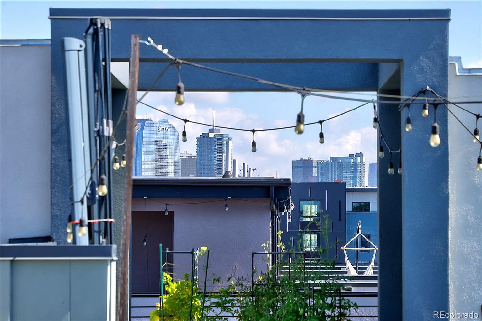 This image showcases a stylish rooftop patio or balcony with a view of the city skyline. String lights add a warm ambiance to the outdoor space, which features modern architectural elements and lush greenery. A hammock chair is visible, suggesting a relaxing and inviting atmosphere.