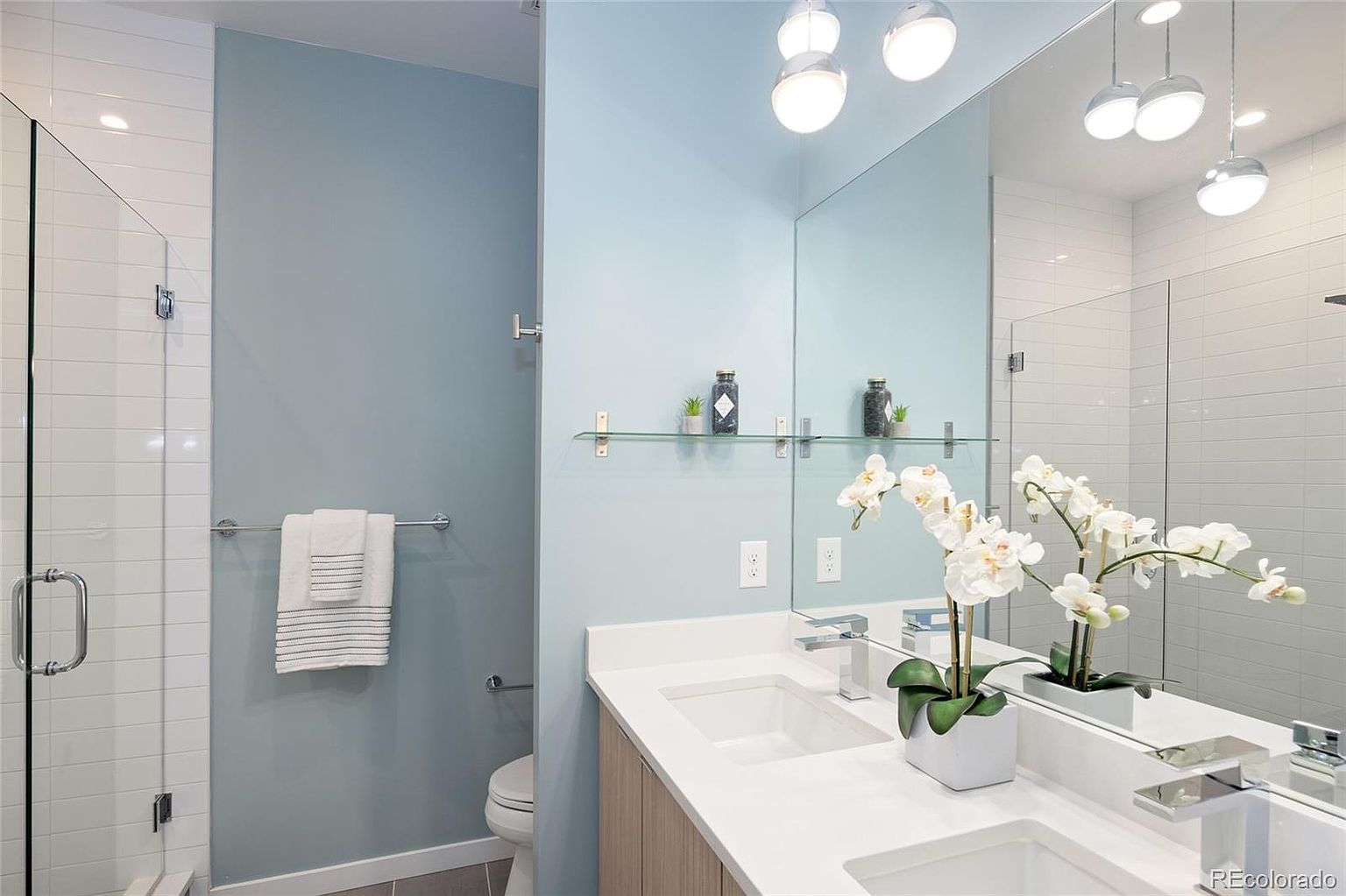 This is a well-lit bathroom featuring a double vanity with white countertops and light wood cabinetry. A large mirror reflects the vanity area and the modern lighting fixtures above. The walls are painted in a soft blue hue, and a glass-enclosed shower is visible to the left, creating a clean and contemporary aesthetic.