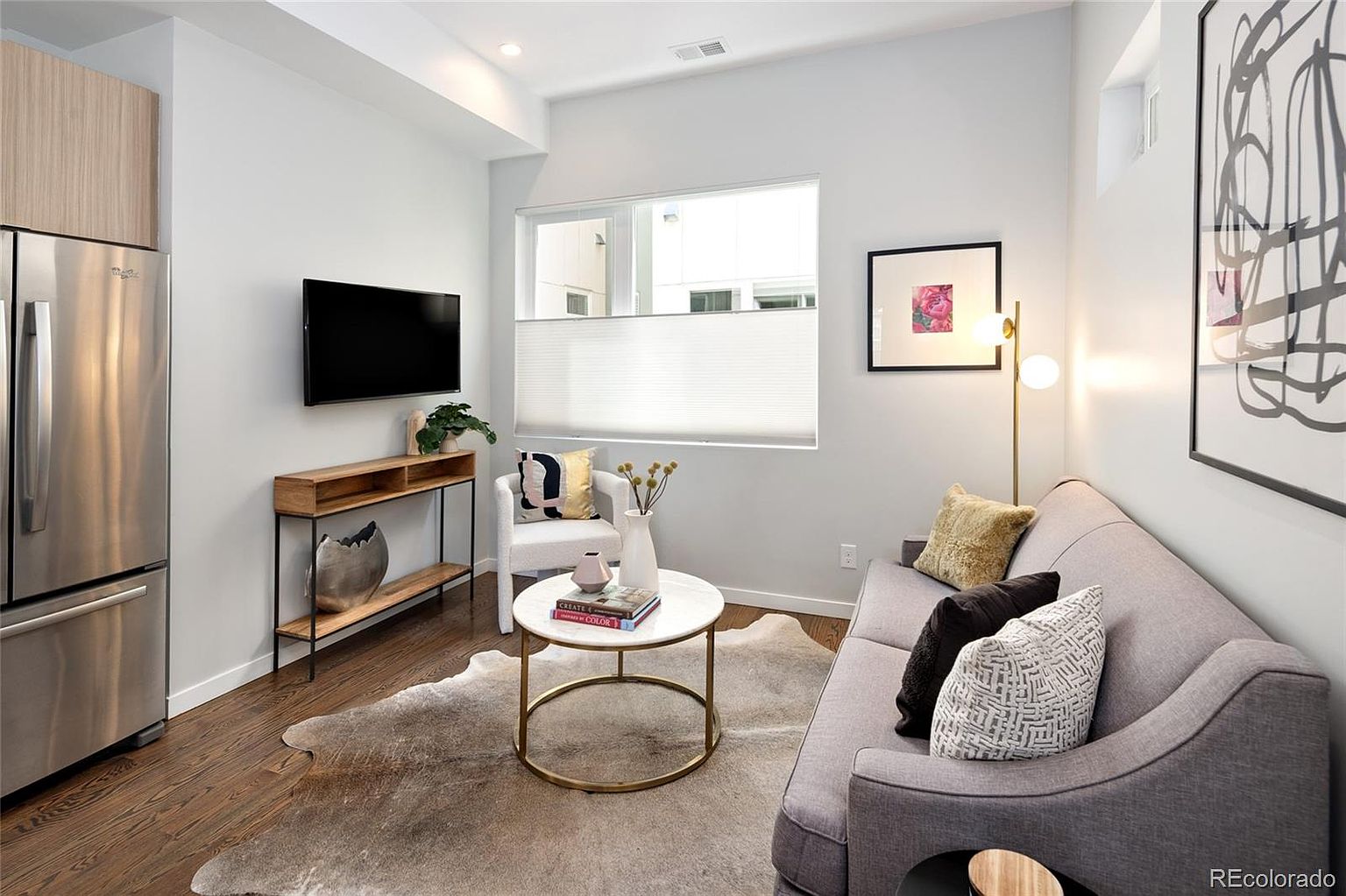 This is a cozy living room featuring a gray sofa with decorative pillows, a round coffee table, and a console table with a TV mounted above. A cowhide rug adds texture to the hardwood floors, and natural light streams in through a window with blinds. The overall impression is modern and inviting.