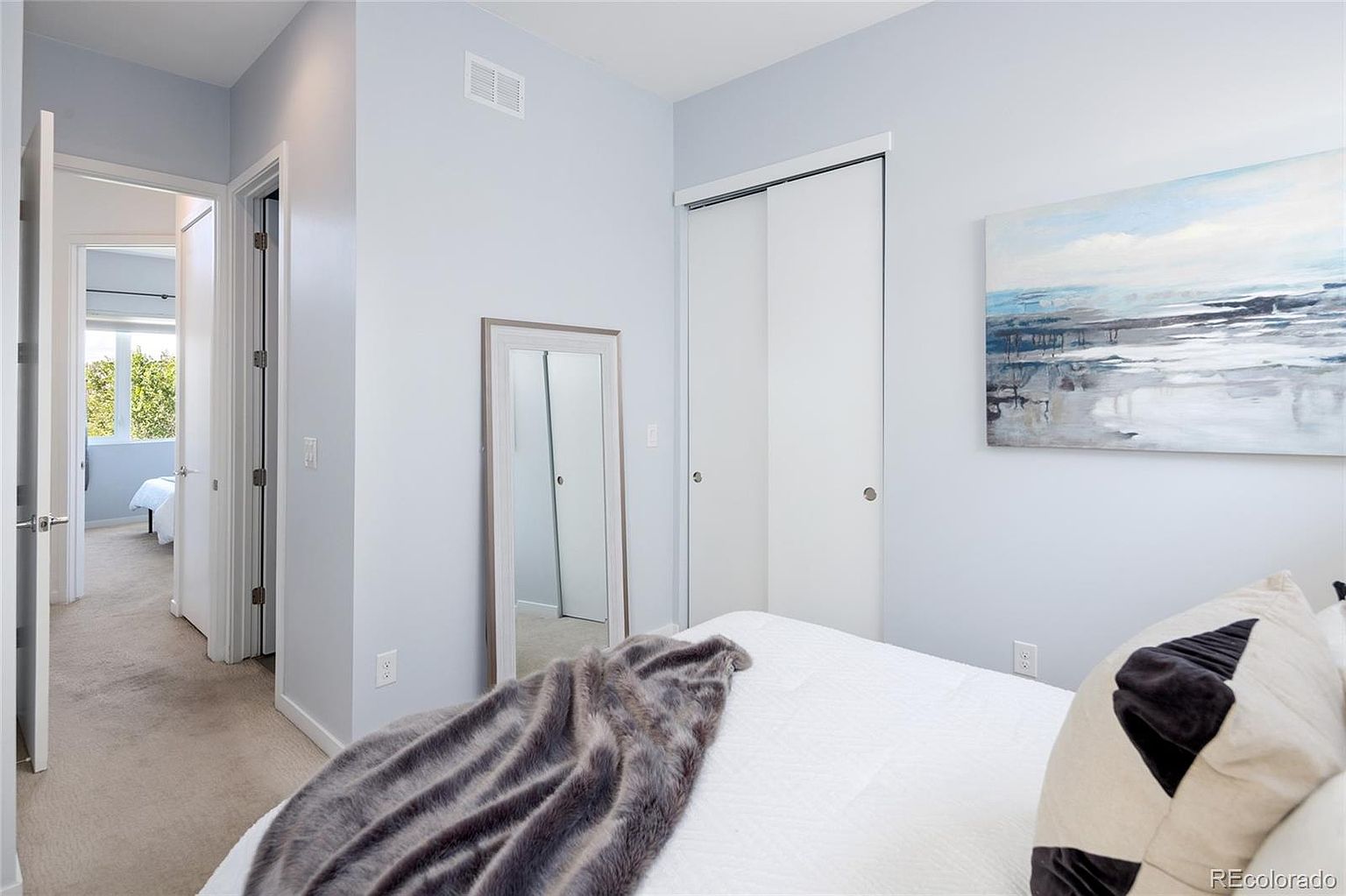 This is a bright and airy bedroom featuring light blue walls, a white bed with decorative pillows and a faux fur throw. The room includes a full-length mirror, a closet with sliding doors, and a modern abstract painting. The space is clean and well-lit, creating a calming and inviting atmosphere.