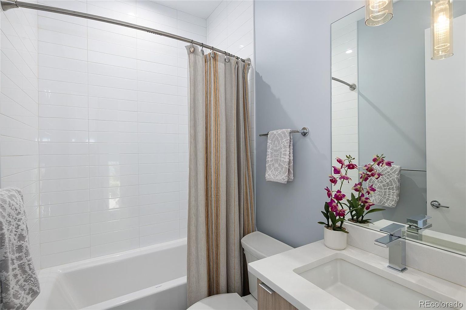 This is a well-lit bathroom featuring a white tiled shower/tub combination with a neutral-toned shower curtain. The vanity has a white countertop, a modern faucet, and a decorative orchid. The walls are painted a soft blue, and a towel rack holds a patterned towel, creating a clean and inviting space.