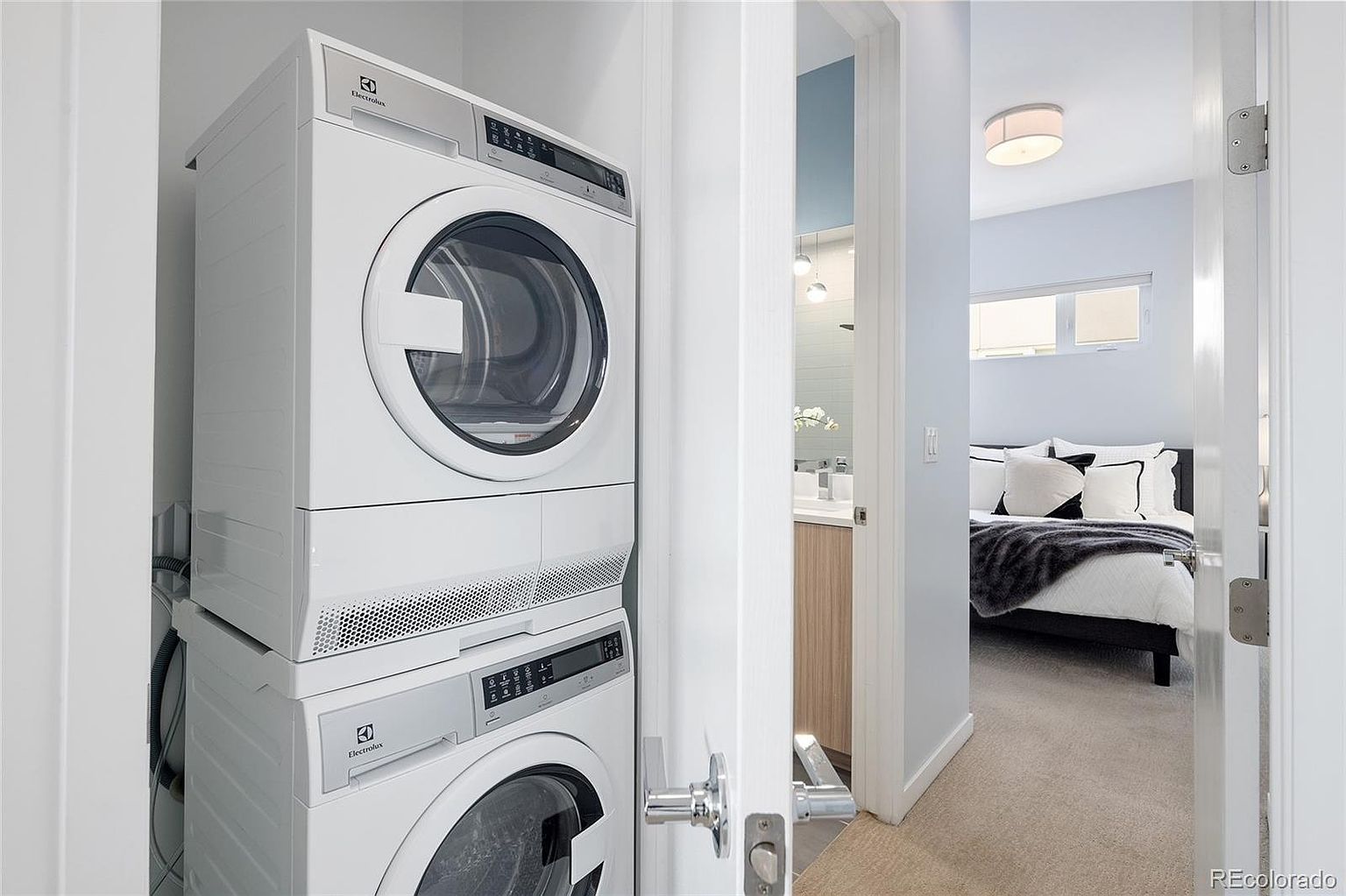 The image showcases a laundry area with a stacked Electrolux washer and dryer set. The laundry space is adjacent to a hallway leading to a bedroom and bathroom, visible through open doorways. The overall impression is clean and modern, highlighting the convenience of in-unit laundry.