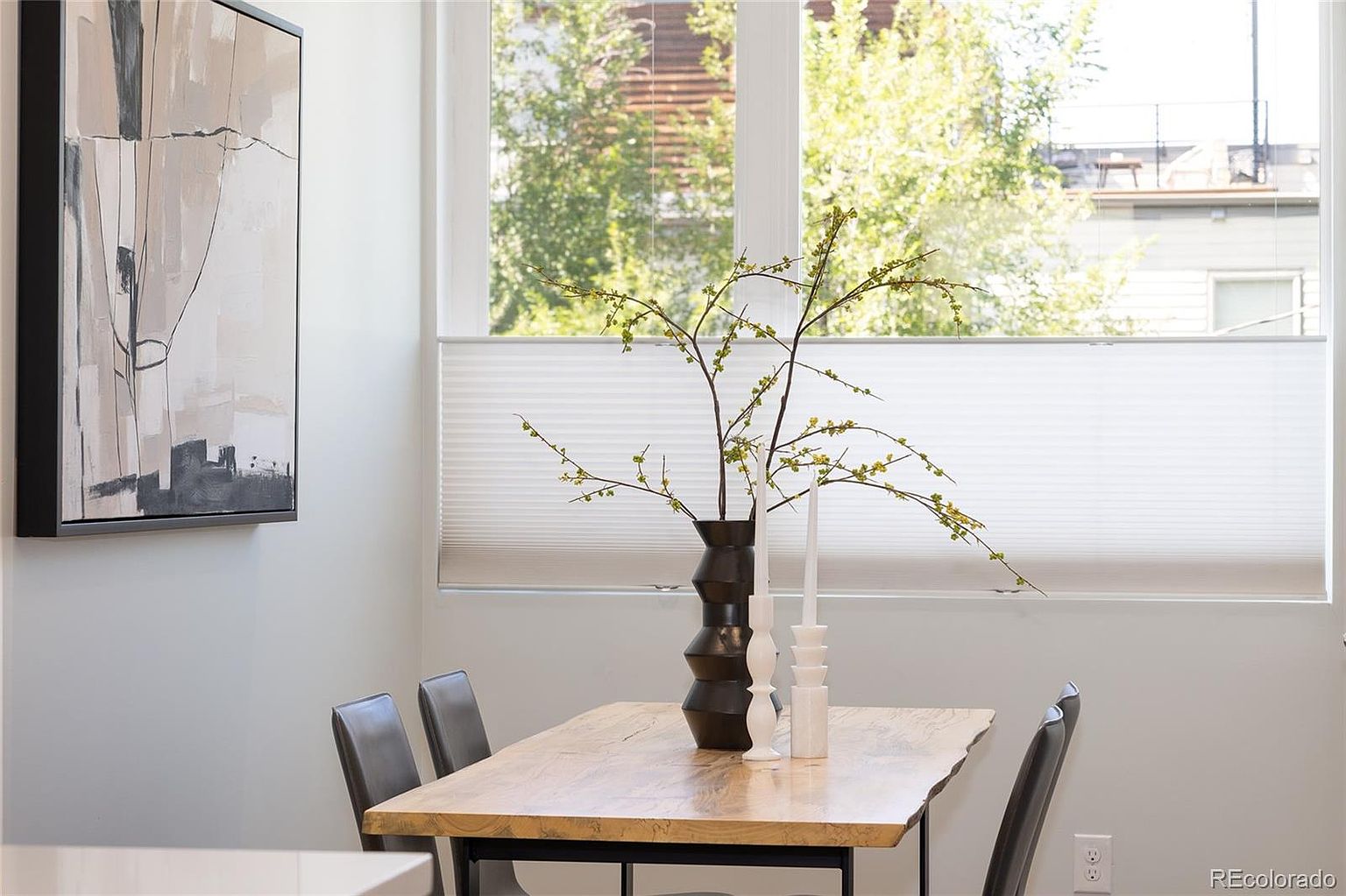 This is a dining room featuring a wooden table with a black metal frame, surrounded by modern chairs. A decorative vase with branches and candles sits on the table, adding a touch of elegance. A large window with blinds provides natural light, and an abstract painting hangs on the wall, enhancing the room's aesthetic appeal.