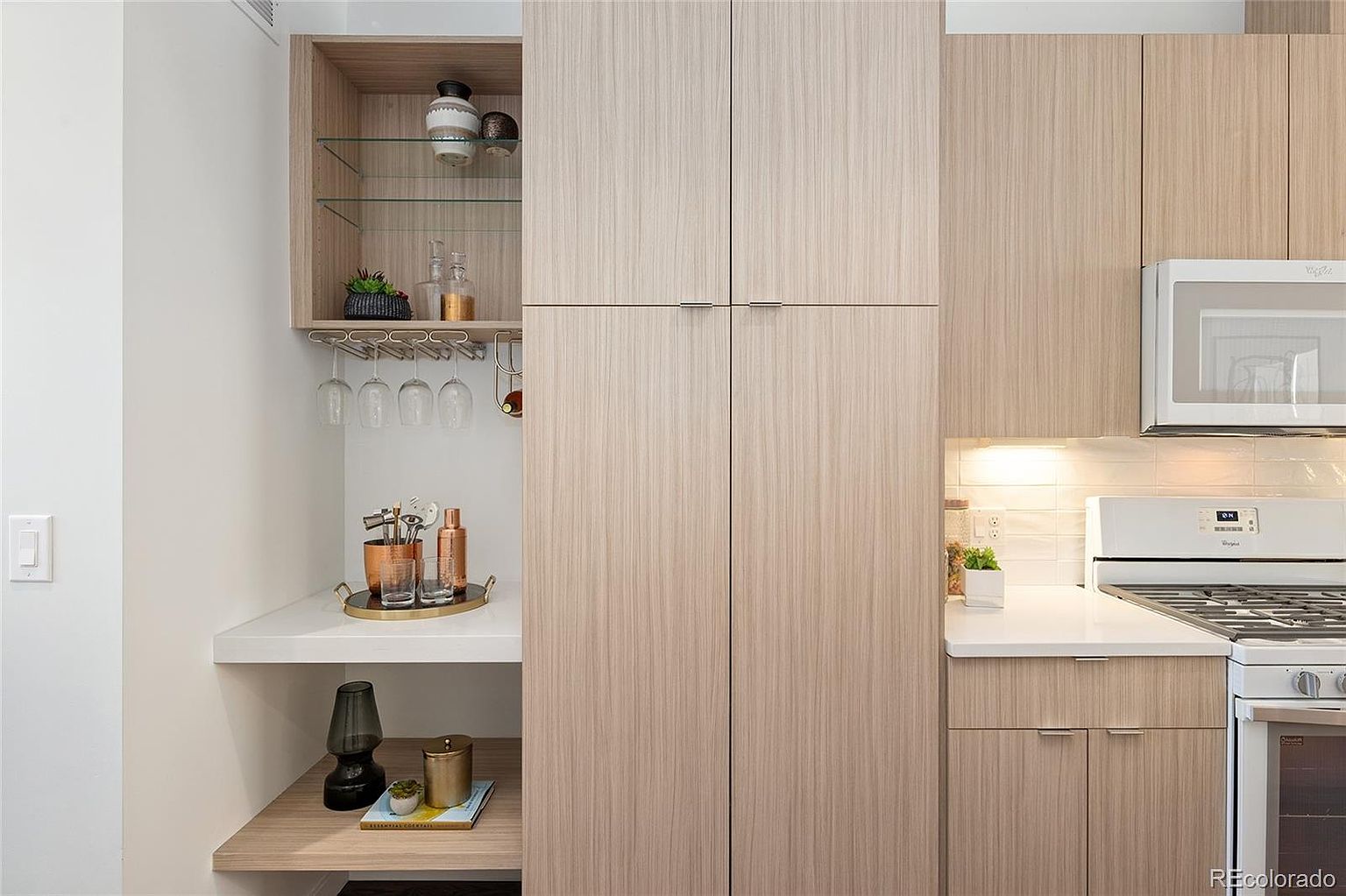 This is a well-organized kitchen featuring light wood cabinetry and white countertops. The space includes a built-in bar area with glass shelves and wine glass storage, as well as modern appliances such as a microwave and gas range. The perspective is a medium shot, highlighting the functionality and style of the kitchen.