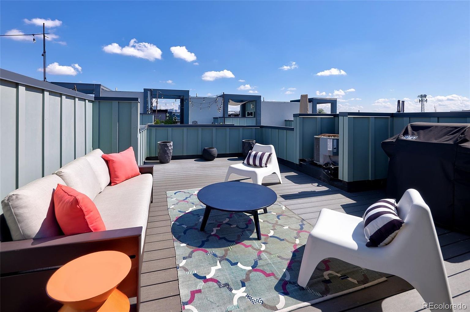 This image showcases a stylish rooftop patio featuring modern outdoor furniture, including a sofa with coral pillows, two white chairs with a striped pillow, and a round blue coffee table on a patterned rug. The deck is surrounded by a light blue privacy fence, offering a secluded and inviting outdoor living space with a grill on the side. The sky is blue with some clouds.