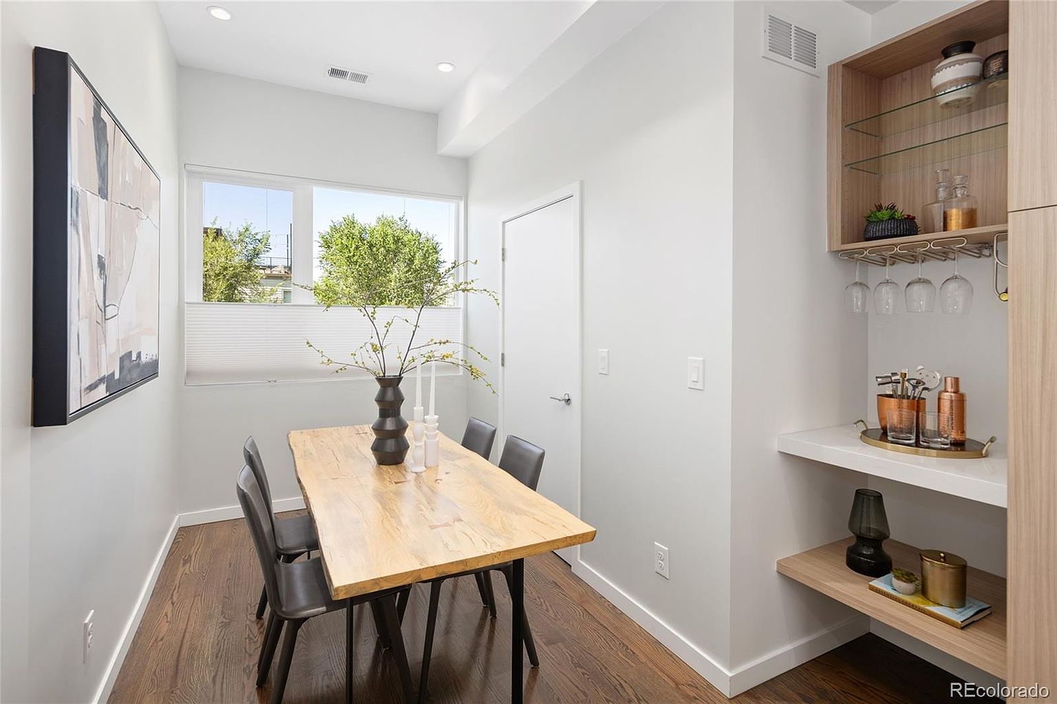 This is an interior shot of a dining room featuring a wooden table with black legs and gray chairs. A black vase with branches and white candles sits on the table. A built-in bar area with shelves and wine glass rack is on the right side of the room. The room has hardwood floors and white walls, with a modern painting on the left wall.