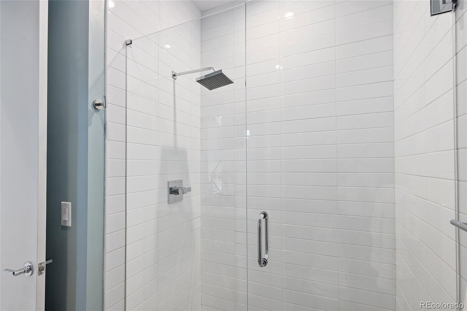 This is a well-lit primary bathroom featuring a modern shower with white, horizontally-laid tiles and a frameless glass enclosure. The shower includes a square showerhead and sleek chrome fixtures. The overall impression is clean, contemporary, and luxurious, highlighting the bathroom's modern design.