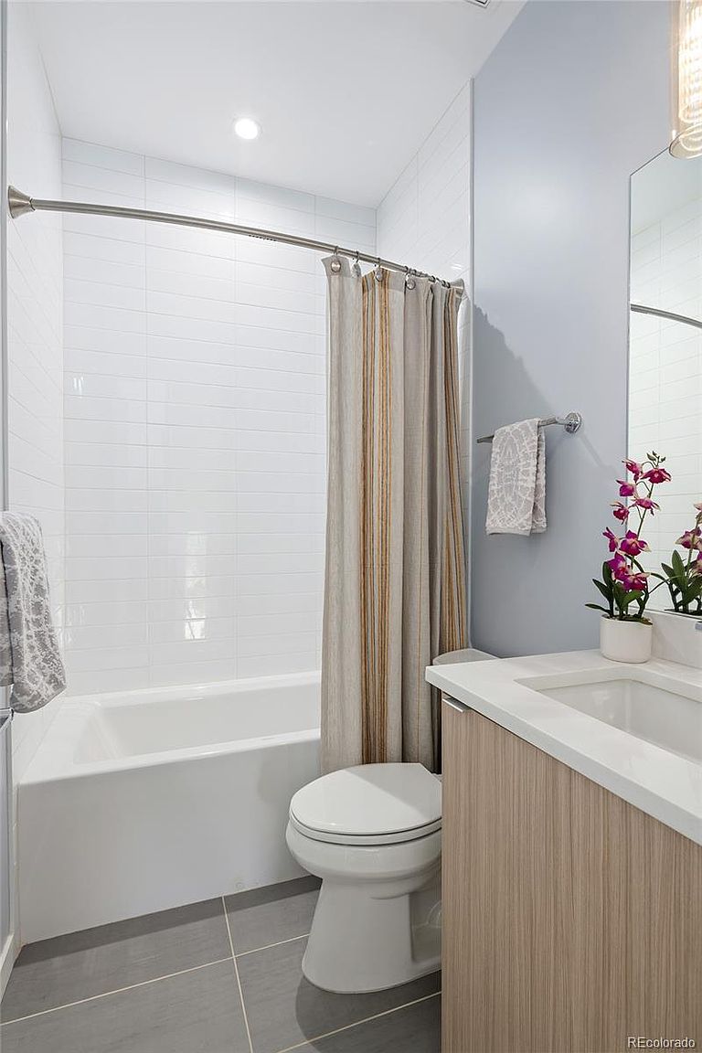This is a well-lit bathroom featuring a bathtub with white tiled walls and a shower curtain. A toilet is positioned next to the tub, and a vanity with a white countertop and light wood-grain cabinetry is visible. The floor is tiled in a neutral gray, creating a clean and modern aesthetic.