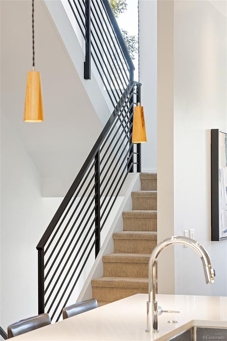This interior shot showcases a modern staircase with black metal railings and carpeted steps. Two pendant lights with wooden shades hang from the ceiling, adding a warm touch to the otherwise white and neutral color scheme. The view is from the bottom of the stairs, looking up, with a partial view of a kitchen counter and sink in the foreground.