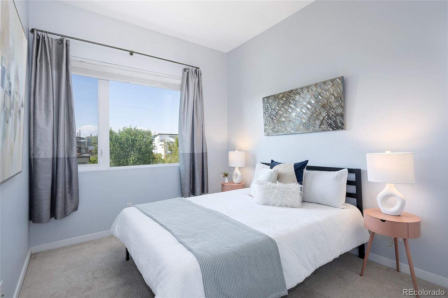 This is a cozy bedroom featuring a bed with white linens and a light blue throw, flanked by matching pink nightstands and lamps. The room has a modern aesthetic with light blue walls, gray curtains, and a decorative metal artwork above the bed. A window provides natural light and a view of the outdoors.