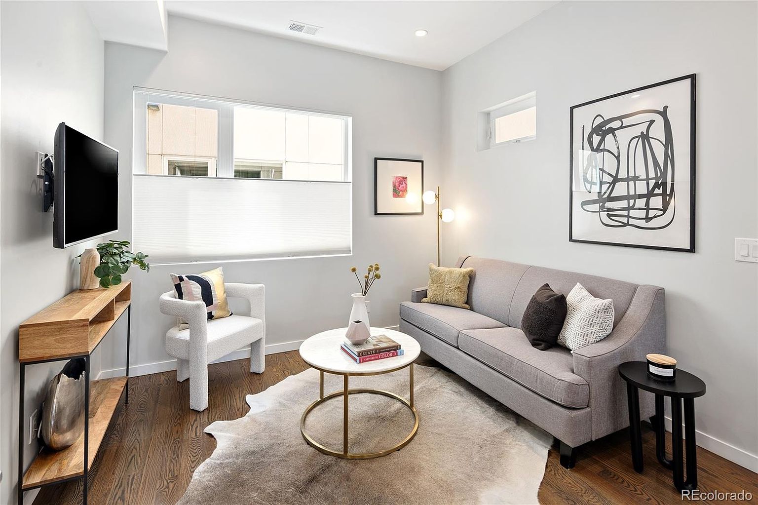 This is a cozy living room featuring a gray sofa with decorative pillows, a white accent chair, and a round coffee table on a cowhide rug. A modern console table sits beneath a wall-mounted TV, and abstract art hangs on the wall, creating a stylish and inviting atmosphere. The room is well-lit with natural light from a window and a floor lamp.