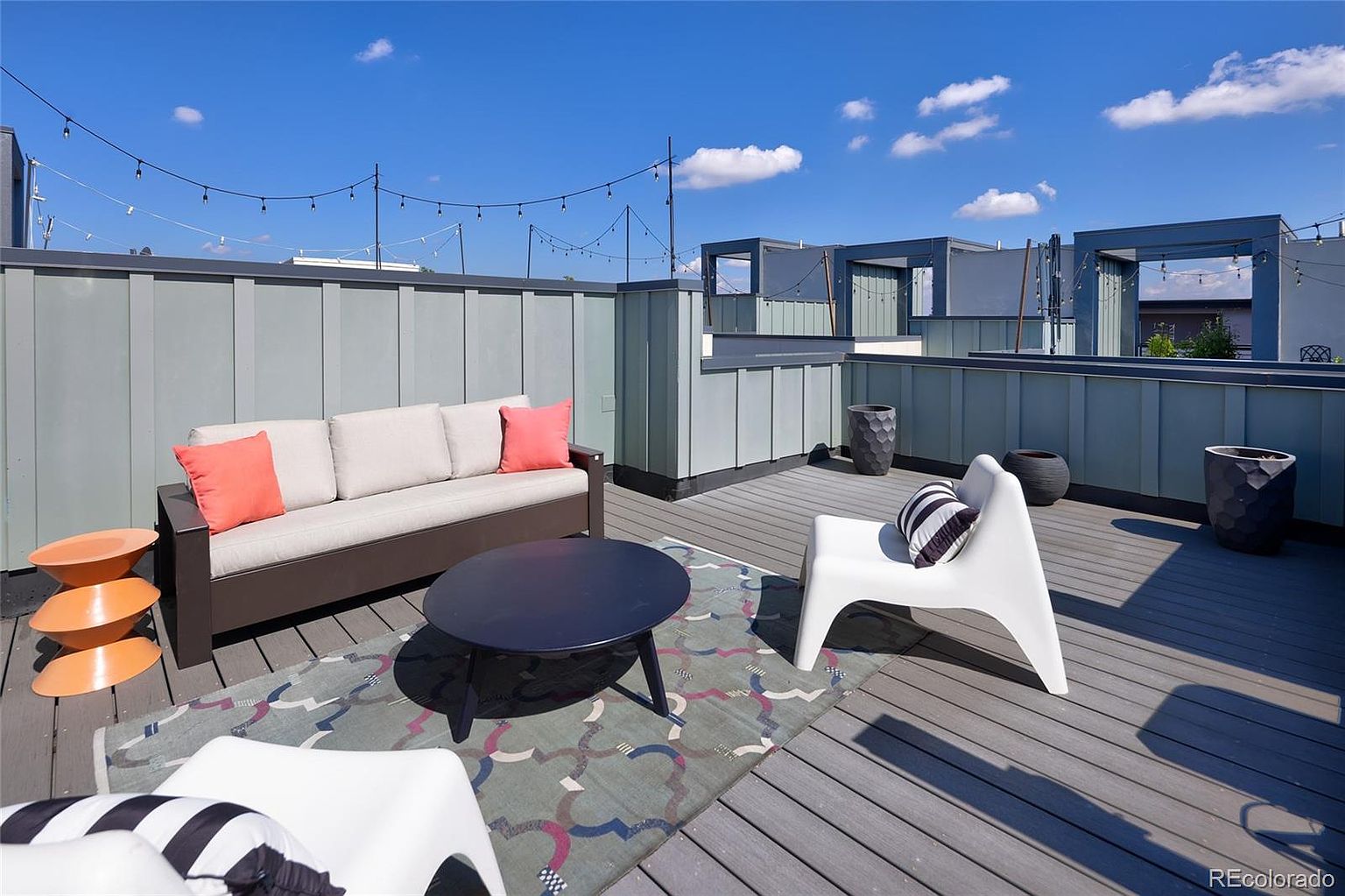 This image showcases a stylish rooftop patio or deck area. The space is furnished with a modern sofa, chairs, and a coffee table, arranged on a patterned rug. The deck features a gray wood-like surface, and the surrounding walls are a muted green, creating a contemporary and inviting outdoor living space.