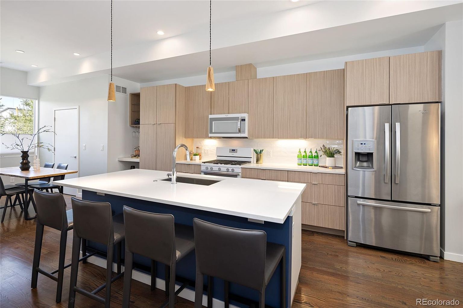 This is a well-lit, modern kitchen featuring light wood cabinetry and stainless steel appliances, including a large refrigerator. A central island with a white countertop and blue base provides ample workspace and seating with four bar stools. The kitchen also includes pendant lighting and a view into the dining area, creating an open and inviting space.