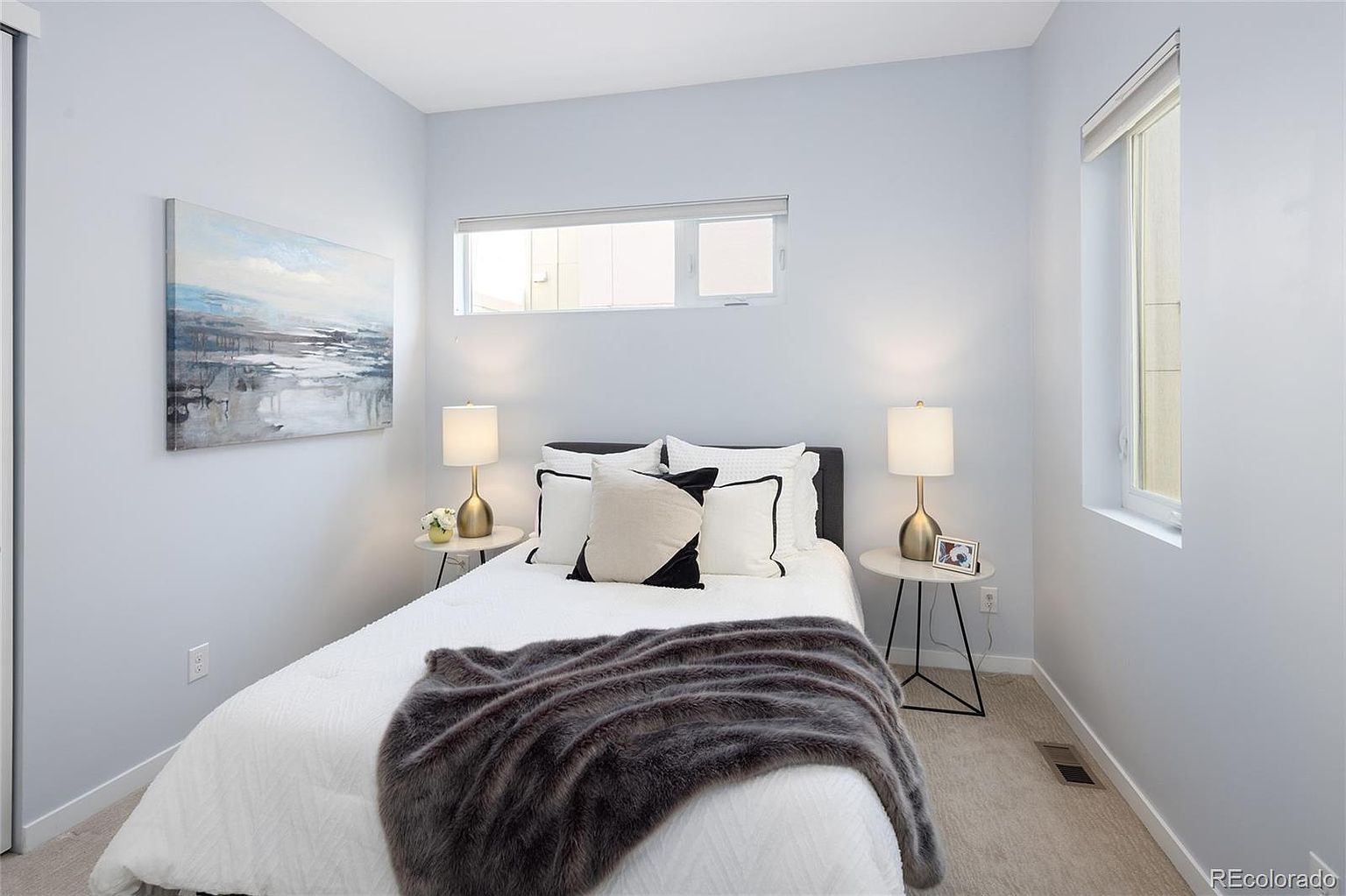 This is a cozy bedroom featuring a bed with white linens, black and white decorative pillows, and a gray faux fur throw. Two bedside tables with gold lamps flank the bed, and a modern art piece hangs on the wall. A horizontal window provides natural light, creating a serene and inviting atmosphere.
