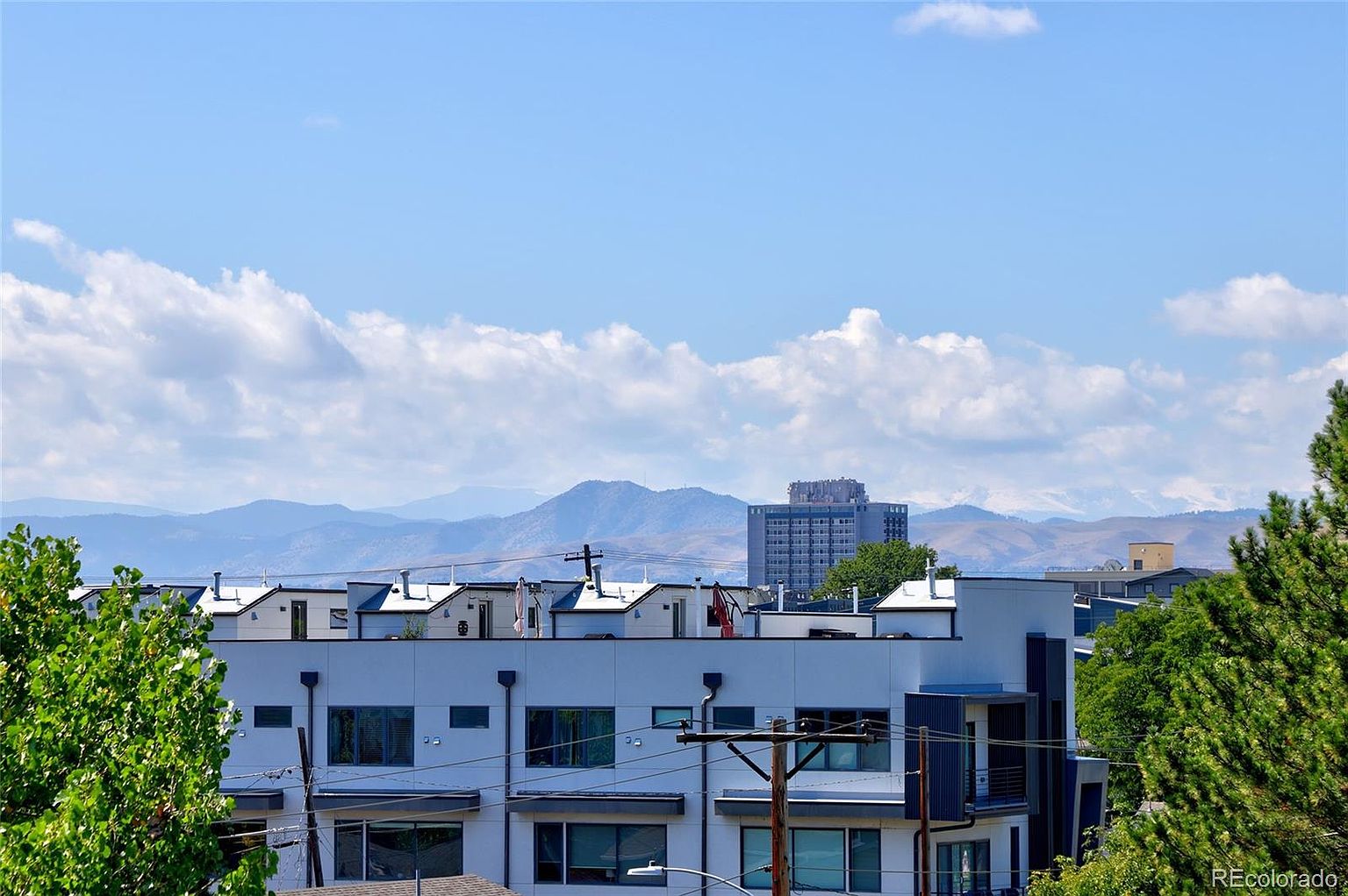 This image showcases the front view of a modern townhouse complex under a bright, partly cloudy sky. The buildings feature a contemporary design with clean lines, flat roofs, and a neutral color palette. Mountains are visible in the background, adding depth and scenic appeal to the property's location.