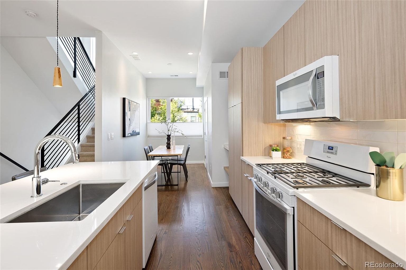 This is a well-lit, modern kitchen featuring light wood cabinetry and white countertops. Stainless steel appliances, including a range and microwave, are seamlessly integrated. The kitchen flows into a dining area with a view to the outside, and a staircase is visible on the left, creating an open and inviting space.