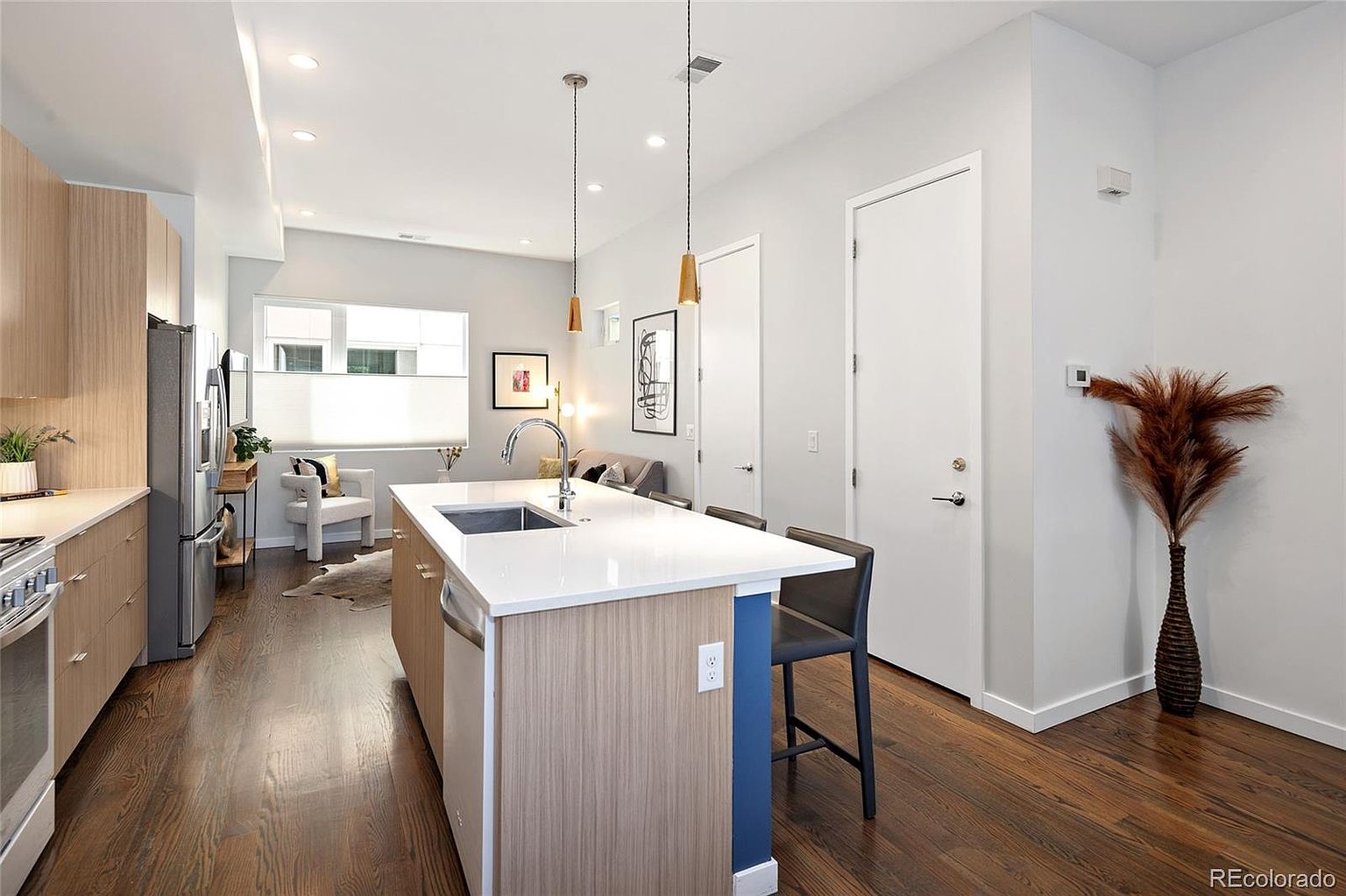 This is an interior shot of a modern kitchen with hardwood floors and light gray walls. The kitchen features a central island with a white countertop, light wood cabinetry, and a stainless steel sink. Pendant lights hang above the island, and stainless steel appliances are visible along the left wall. A vase with decorative branches sits against the wall near a white door, adding a touch of elegance.