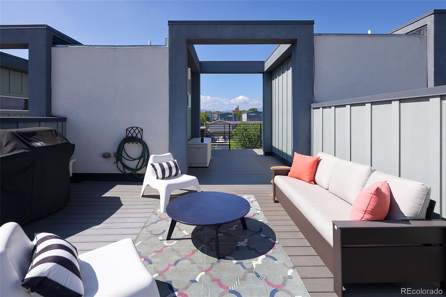 This image showcases a stylish rooftop deck or patio area. The space is furnished with a comfortable sofa, chairs, and a round coffee table, all arranged on a modern outdoor rug. A grill is visible on the side, suggesting an area for outdoor cooking and entertaining, with a view of the neighborhood in the background.