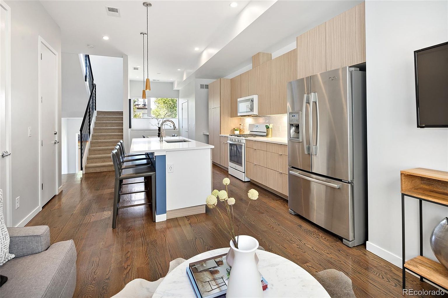 This is an interior shot of a modern kitchen featuring hardwood floors, stainless steel appliances, and light wood cabinetry. A white kitchen island with bar seating is centrally located, and pendant lights hang above it. The space is well-lit and appears to be part of an open-concept living area, with a staircase visible in the background.