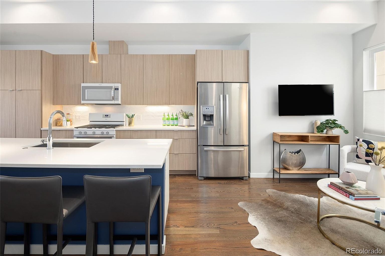 This is a well-lit, modern kitchen and living area. The kitchen features light wood cabinetry, stainless steel appliances, and a white countertop island with seating. The living area includes a TV mounted on the wall, a console table, and a comfortable seating arrangement, all contributing to a stylish and inviting open-concept space.