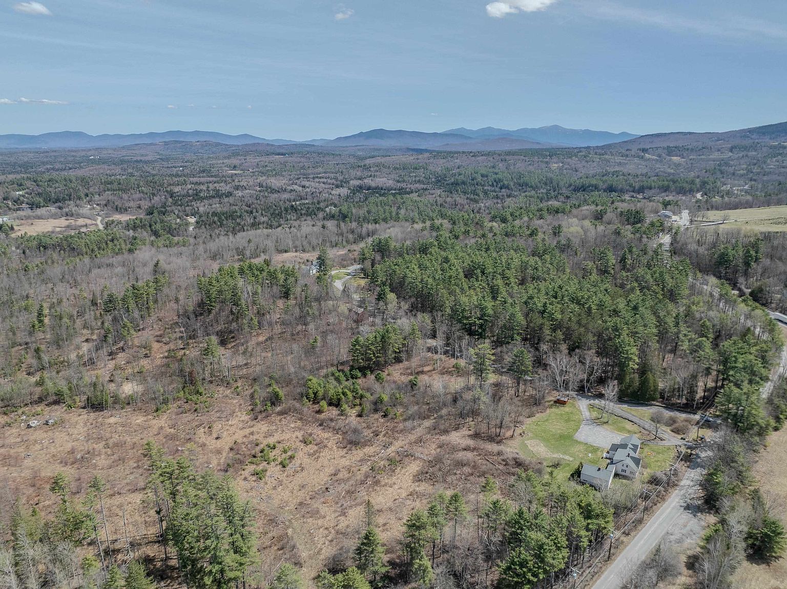 This high-angle aerial perspective captures a rural property nestled within a vast, forested landscape under a clear blue sky. The scene highlights a single-family home with a paved driveway, surrounded by a mix of open fields and dense woodland, with distant mountain ranges providing a scenic backdrop. The composition emphasizes the expansive nature of the lot and its quiet, secluded setting.