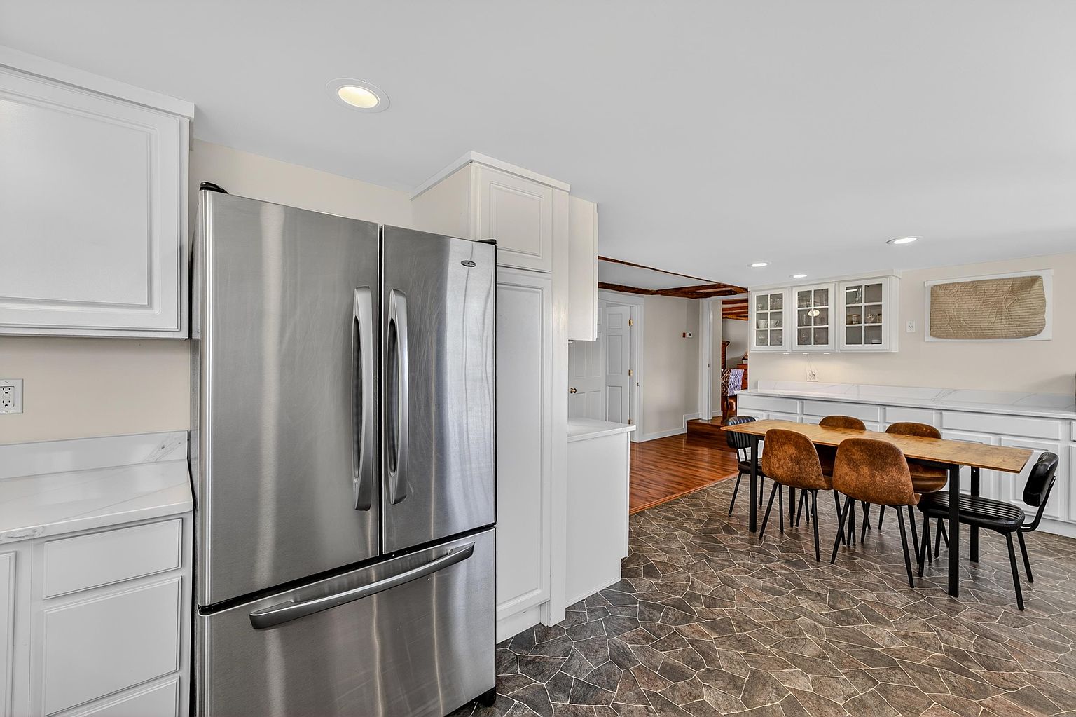 This kitchen features white cabinetry paired with a stainless steel refrigerator and a long dining table set for several guests. The space is anchored by a distinctive stone-patterned tile floor that adds texture to the room, while a transition leads into a hallway with warm-toned wood flooring. The perspective captures the open-concept layout, highlighting the functional flow between the food preparation area and the adjacent dining space.