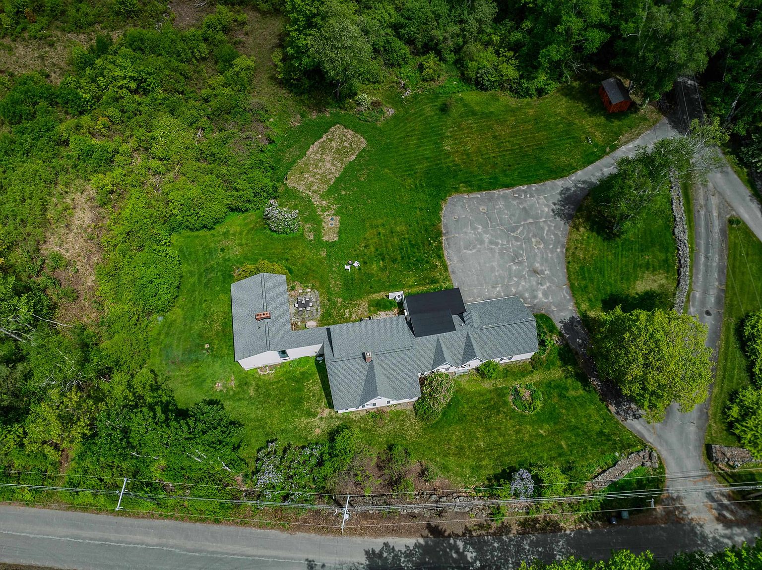 This high-angle aerial perspective captures a sprawling residential property nestled within a lush, wooded landscape. The home features a multi-gabled roofline with grey shingles, a paved driveway leading to a parking area, and a private backyard with a stone patio. The surrounding grounds include expansive green lawns, mature trees, and a small outbuilding, offering a sense of seclusion and natural beauty.