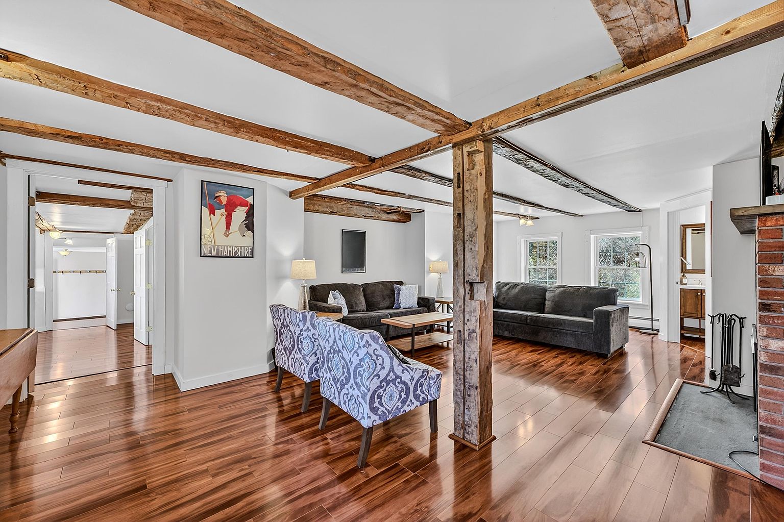 This spacious living room features warm, polished wood flooring and exposed timber ceiling beams that add a rustic, historic charm to the space. The room is furnished with two dark sofas and a pair of patterned accent chairs, centered around a wooden coffee table. A prominent, weathered wooden support post stands in the foreground, while a brick fireplace surround is visible on the right, creating a cozy and inviting atmosphere.