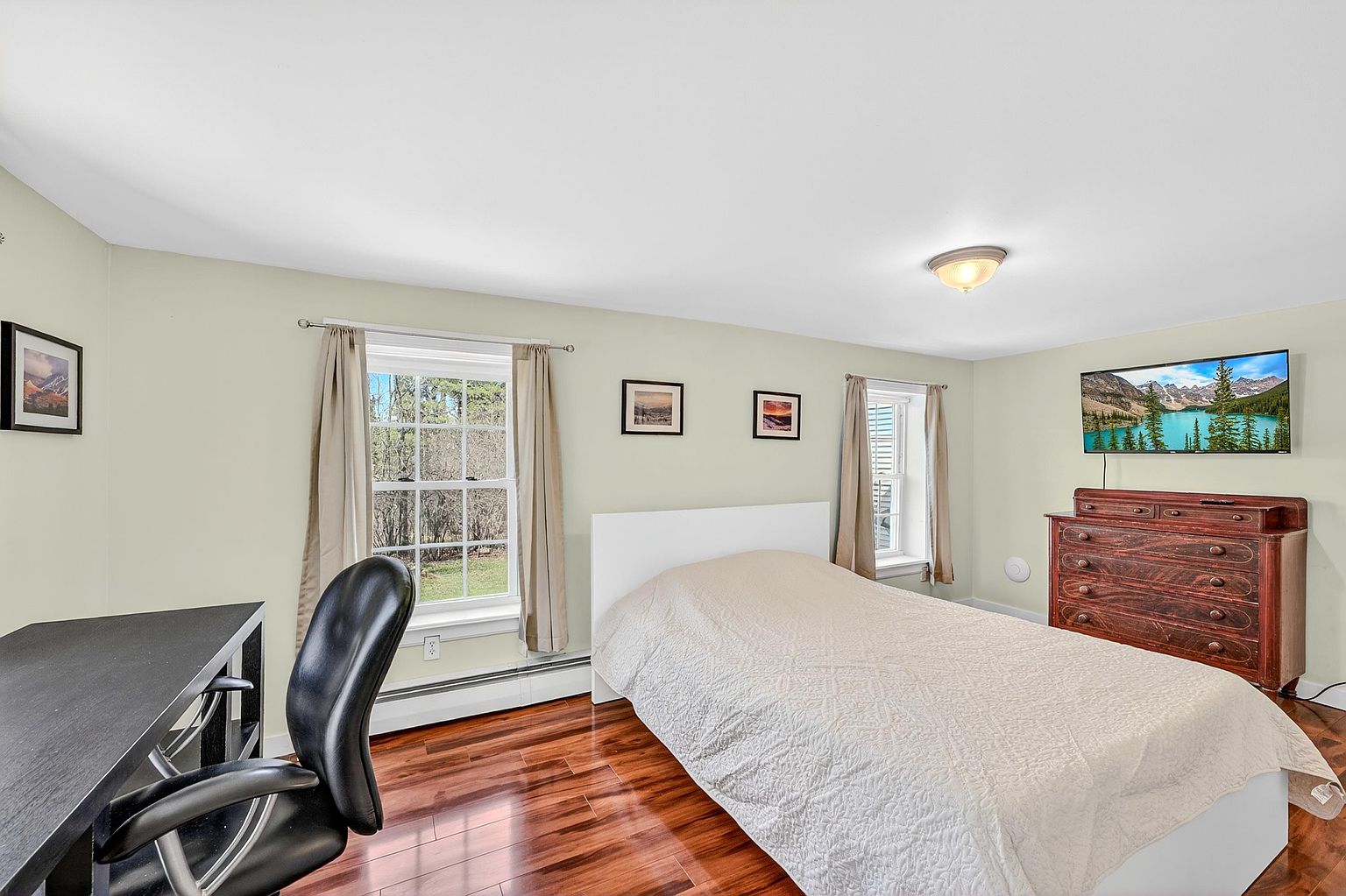 This bedroom features warm-toned wood flooring and light-colored walls, creating a bright and inviting atmosphere. The room is furnished with a bed, a dark wood dresser topped with a television, and a desk with a black office chair, making it a versatile space for sleeping and working. Two windows with neutral curtains allow natural light to fill the room, which is accented by framed artwork on the walls.