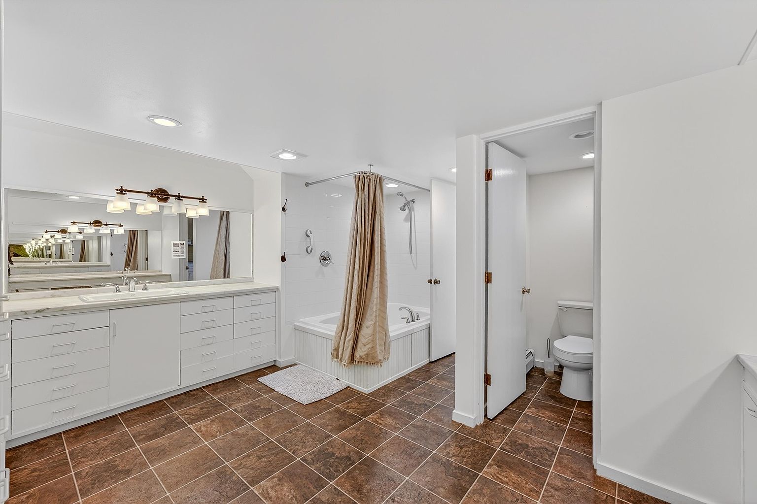 This spacious primary bathroom features a long vanity with ample drawer storage and a large mirror, complemented by a separate tub and shower area. The room is finished with dark, square-patterned floor tiles and white cabinetry, creating a clean and functional aesthetic. A private water closet is tucked behind a door to the right, enhancing the layout's privacy and utility.