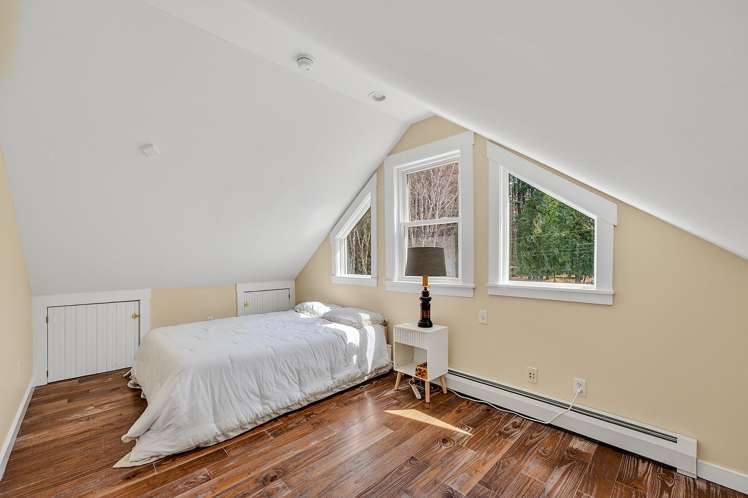 This cozy attic bedroom features sloped ceilings and warm-toned wood-look flooring, creating a comfortable and intimate atmosphere. The space is illuminated by a trio of windows that follow the roofline, providing natural light and views of the surrounding trees. A simple bed and a small bedside table with a lamp complete the minimalist, functional layout of this charming loft space.