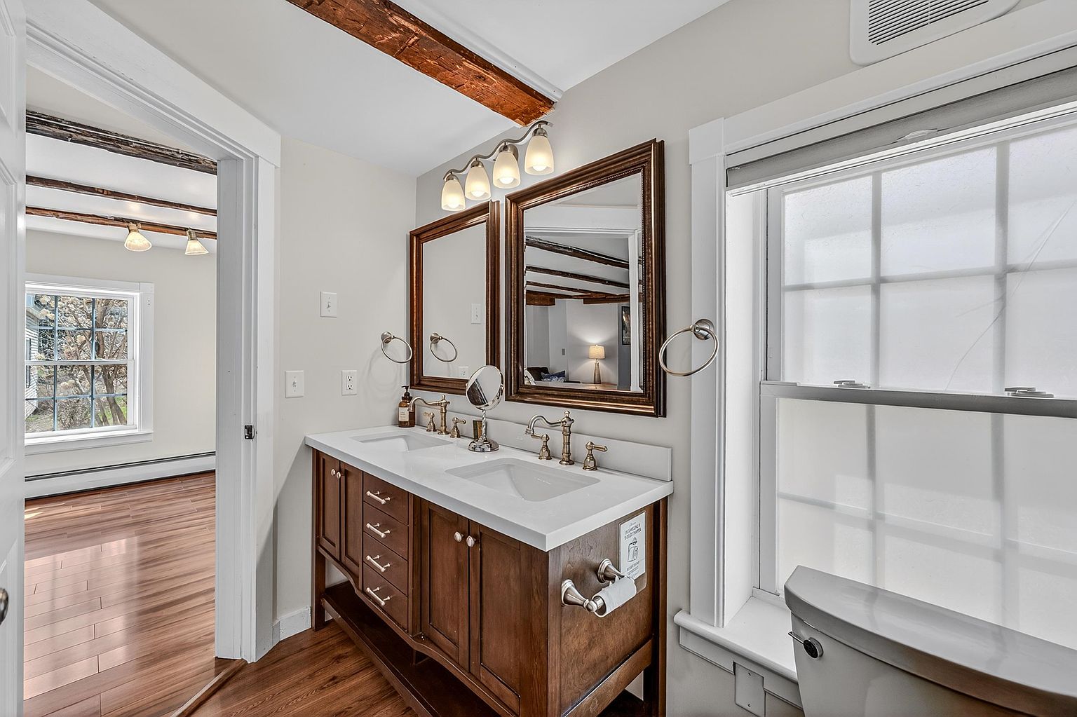 This well-lit bathroom features a double-sink vanity with a dark wood finish and a light-colored countertop, complemented by two framed mirrors and a four-light fixture. The space includes a toilet positioned near a window with frosted glass, and a doorway leads into an adjacent room with wood-look flooring and exposed ceiling beams. The overall aesthetic blends rustic architectural elements with functional, modern bathroom fixtures.