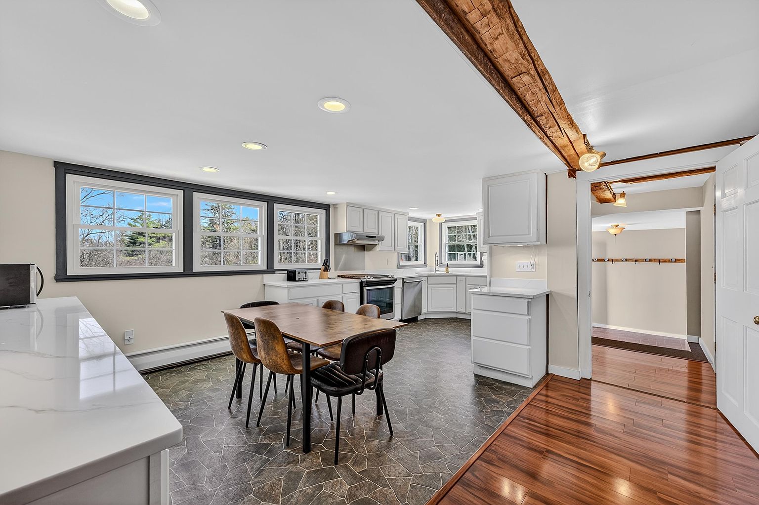 This bright, open-concept kitchen features white cabinetry, stainless steel appliances, and a central dining area with a wooden table and four chairs. The space is defined by a unique combination of dark, geometric-patterned flooring in the kitchen area and warm-toned wood flooring in the adjacent hallway, separated by a rustic exposed wooden ceiling beam. Large windows with dark frames provide ample natural light, creating an inviting and functional atmosphere.