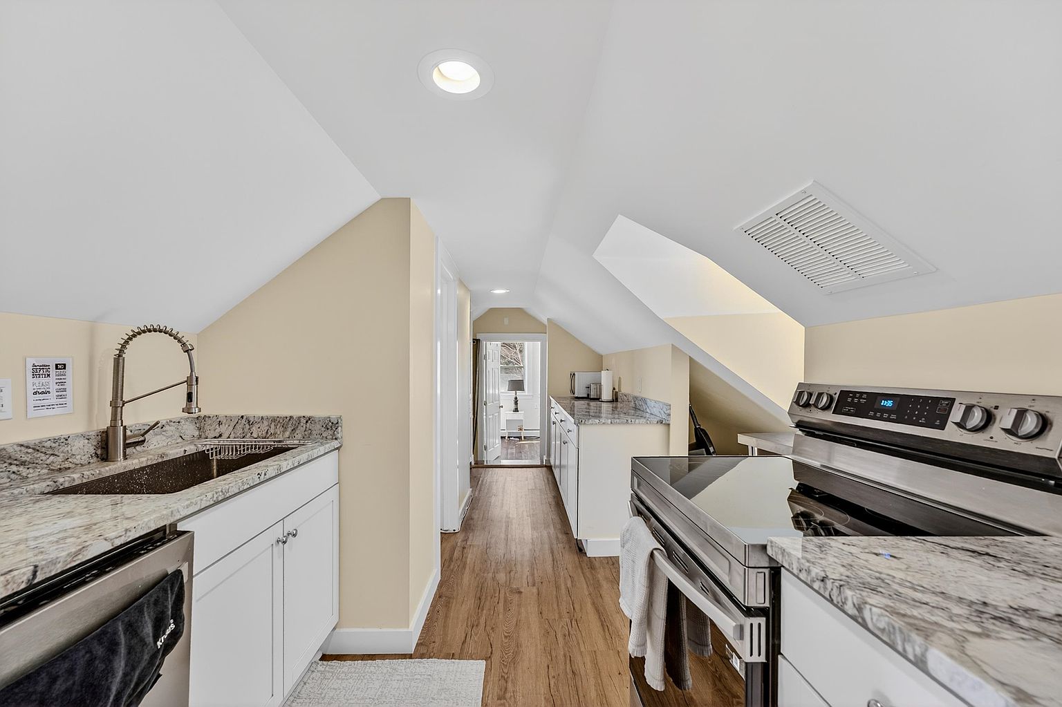 This galley-style kitchen features white cabinetry paired with light-toned stone countertops and wood-look flooring. The space is tucked under a sloped ceiling, creating a cozy, narrow layout that leads to a doorway at the far end. Stainless steel appliances, including a range and a deep sink with a high-arc faucet, provide a functional and modern aesthetic.