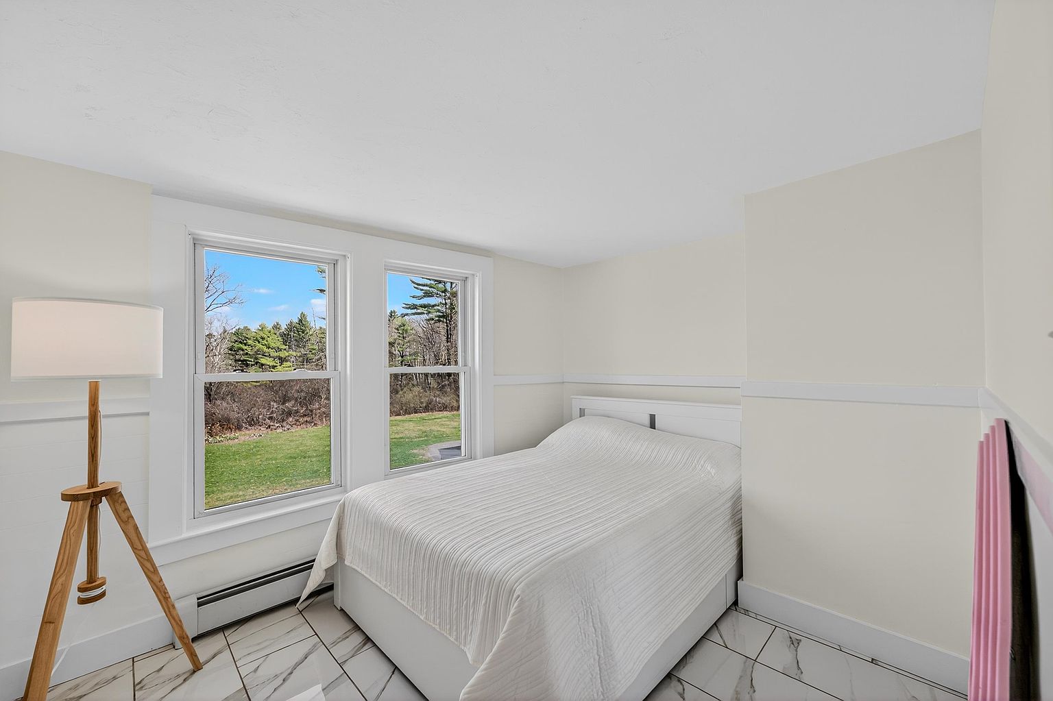 This bright bedroom features a white bed frame with a textured white quilt, positioned against a wall with wainscoting. Two windows provide natural light and a view of the outdoor landscape, while a modern tripod floor lamp stands in the corner. The room is finished with light-colored walls and patterned tile flooring, creating a clean and minimalist aesthetic.