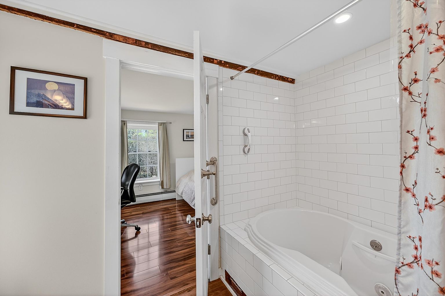 This image captures a view from a bathroom into an adjacent bedroom, highlighting a deep soaking tub surrounded by white subway tile. The bathroom features a classic door with vintage-style hardware, while the bedroom beyond showcases warm wood flooring and a window that brings in natural light. The perspective creates a sense of flow between the private living spaces, emphasizing the home's bright and airy atmosphere.