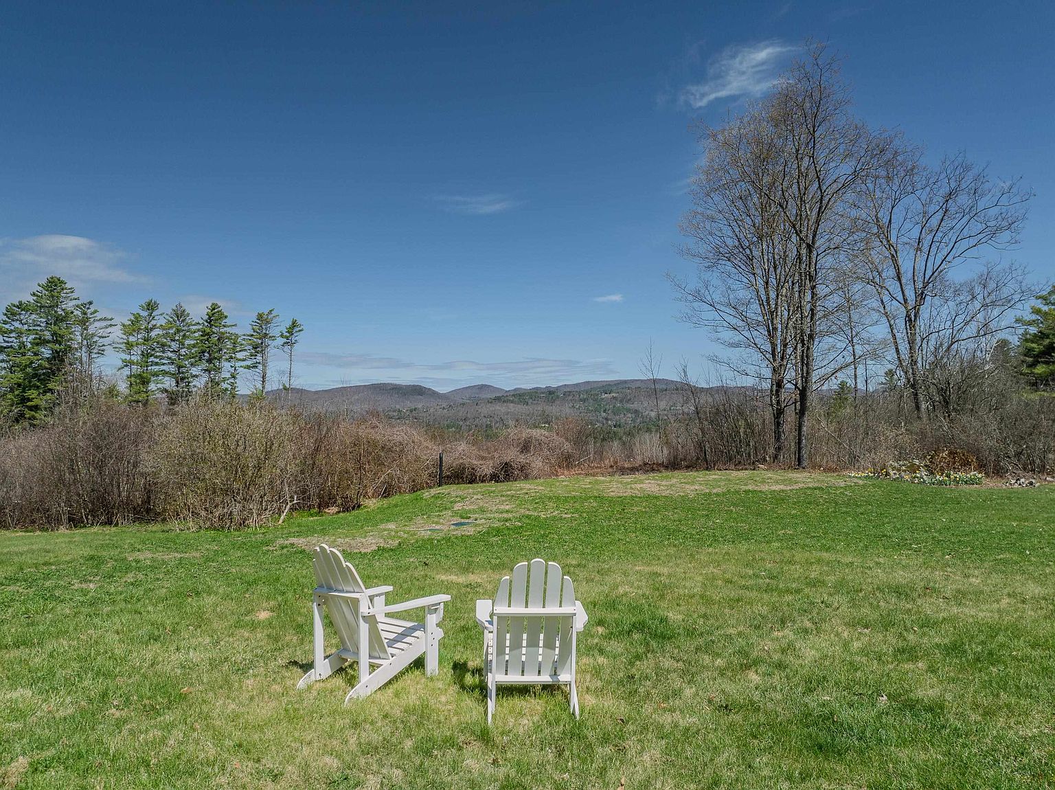 This expansive, grassy backyard offers a serene setting with two white Adirondack chairs positioned to overlook a scenic mountain range in the distance. The landscape features a mix of open lawn, mature trees, and natural brush, creating a peaceful and private atmosphere. The clear blue sky and elevated perspective emphasize the property's connection to the surrounding natural beauty.