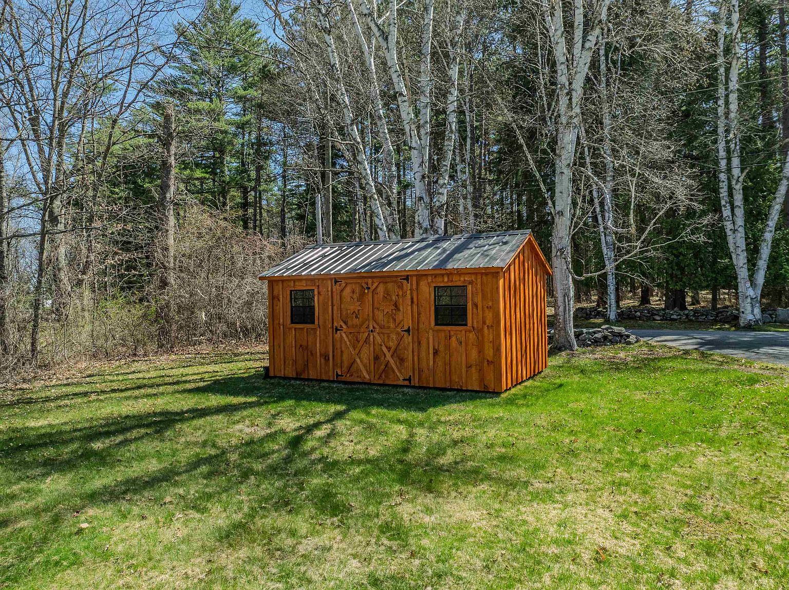 A rustic, wood-sided storage shed sits prominently in the center of a grassy backyard, surrounded by a dense forest of tall, slender trees. The shed features a dark metal roof, double doors with decorative X-bracing, and two small windows, presenting a charming and functional outdoor storage solution. The scene is captured from a slightly elevated perspective, highlighting the shed's integration into the natural, wooded landscape.