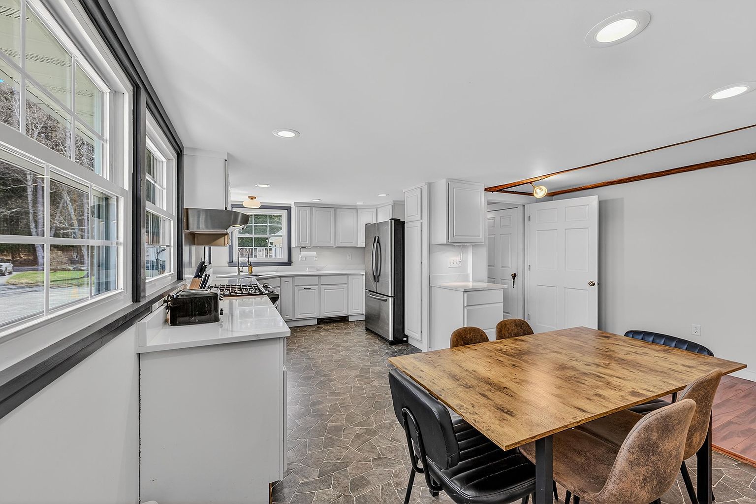 This bright, open-concept kitchen features white cabinetry paired with light-toned countertops and stainless steel appliances. A dining area with a wooden table and dark chairs sits adjacent to the kitchen, creating a functional space for meals. The room is finished with patterned tile flooring and recessed lighting, offering a clean and practical layout.