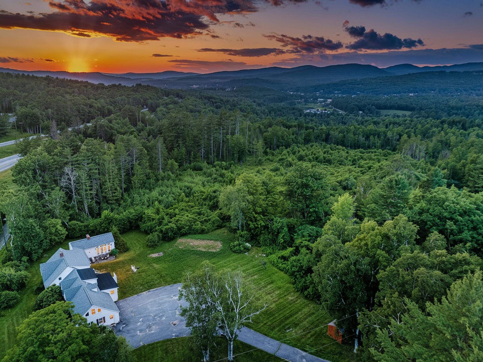 This high-angle aerial shot captures a charming multi-sectioned home nestled within a lush, forested landscape at sunset. The property features a paved driveway leading to the house, surrounded by expansive green lawns and dense woodland that stretches toward distant, rolling mountains under a vibrant, colorful sky. The perspective provides a comprehensive view of the home's layout and its serene, secluded setting.