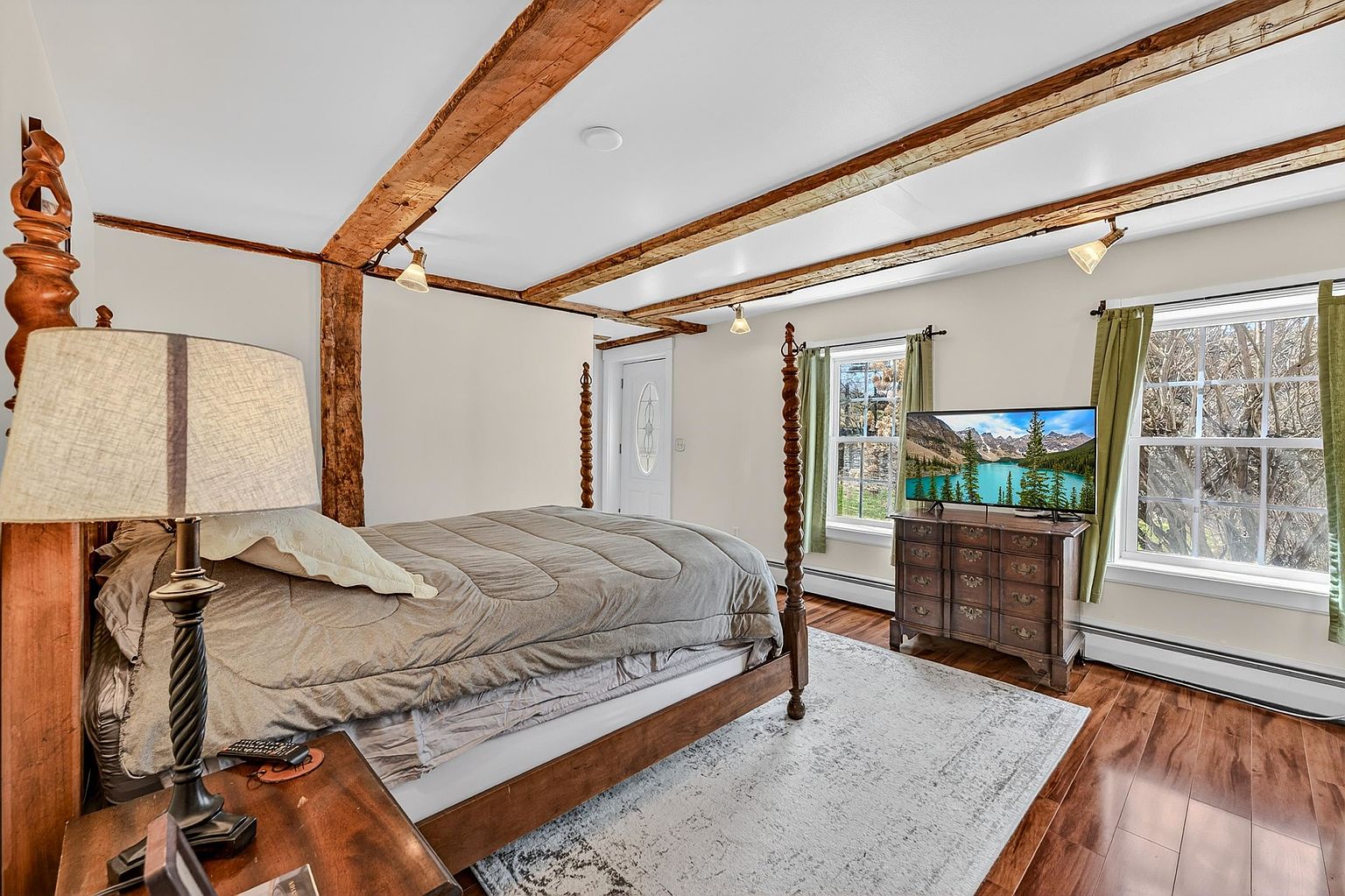 This primary bedroom features rustic exposed wooden ceiling beams and warm-toned wood flooring, creating a cozy, traditional atmosphere. The room is anchored by a four-poster bed with a neutral-toned comforter, complemented by a wooden dresser topped with a television and a bedside lamp. Large windows with green curtains allow natural light to fill the space, while a patterned area rug adds texture to the room.