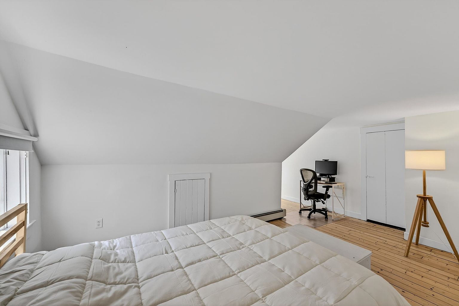 This attic-style bedroom features a sloped ceiling and light wood flooring, creating a cozy and minimalist atmosphere. The room is furnished with a bed covered in a white comforter, a small desk with a computer setup, and a tripod floor lamp. A small access door is built into the knee wall, and the space is illuminated by natural light from a window on the left.