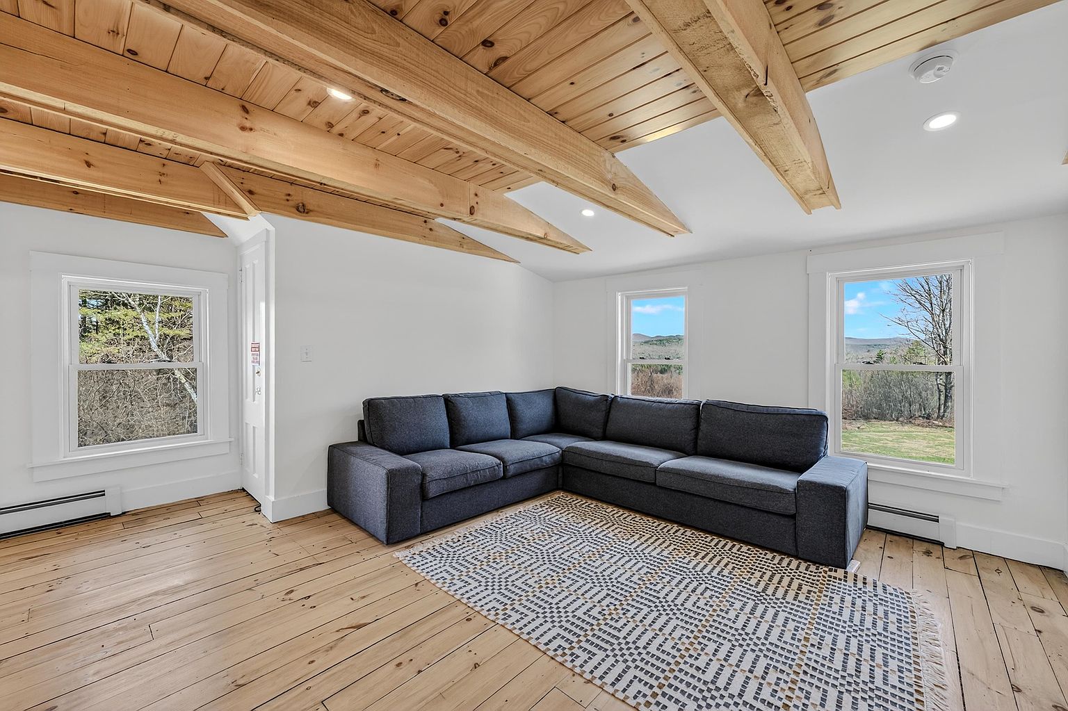 This bright living room features a large, dark gray sectional sofa centered on a patterned area rug atop light-toned wood flooring. The space is defined by its vaulted ceiling with exposed light wood beams and white walls, creating an airy, rustic-modern aesthetic. Three windows provide natural light and views of the surrounding landscape, while recessed lighting adds to the clean, minimalist atmosphere.