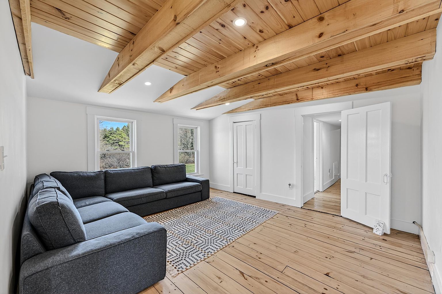 This bright living room features a vaulted ceiling with exposed light wood beams and recessed lighting. A large, dark gray sectional sofa sits atop a patterned area rug, contrasting with the light-toned wood flooring. The space is framed by white walls and two windows that provide natural light, with an open doorway leading to an adjacent hallway.